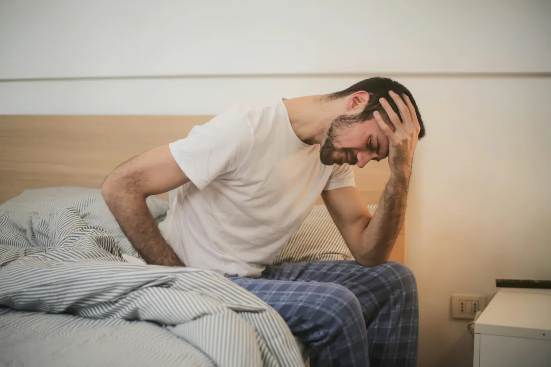 Man sitting on the edge of his bed, holding his lower back and his head in pain after waking