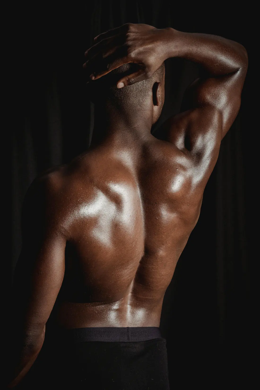 Muscular back of a man with one hand behind his head, photographed against a dark background