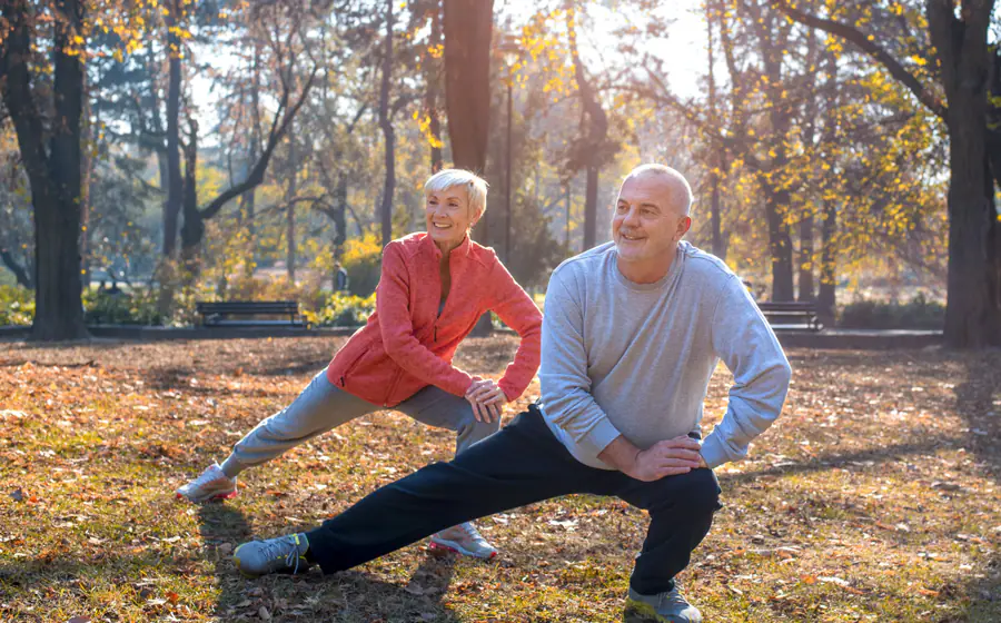 Smiling older couple in sportswear doing side-lunge stretches together in a park on an autumn day