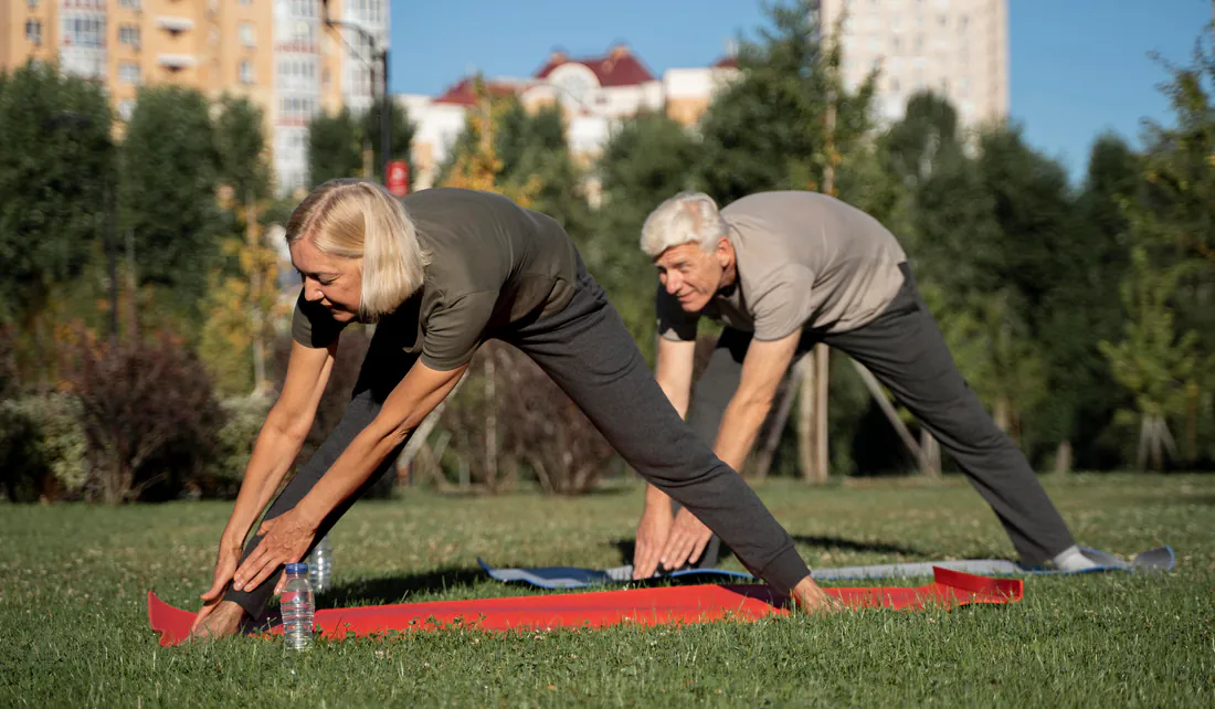 Older couple bending forward to stretch their hamstrings on mats in a sunny park