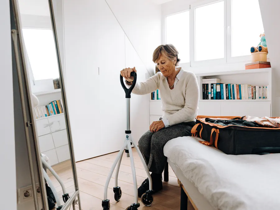 Older woman sitting on her bed beside a four-wheeled rollator walking aid in a bright bedroom